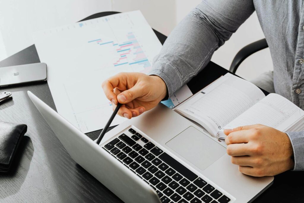 Henkilö analysoi kaavioita kannettavan tietokoneen ja muistikirjan avulla.